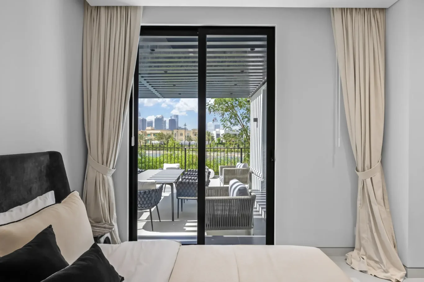 Bedroom view through glass balcony door to an outdoor seating area and distant city skyline carefully framed by beige curtains.