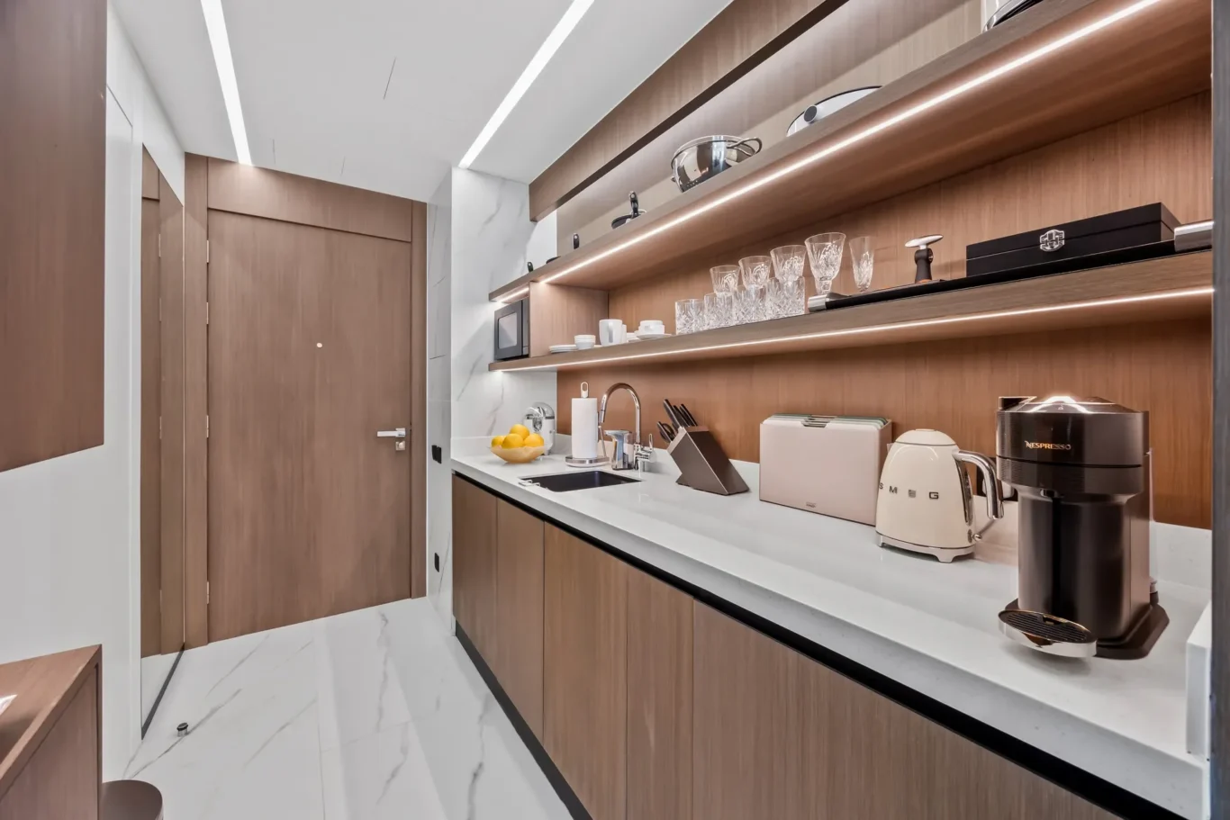 Modern galley kitchen with wood-tone cabinets, white countertop, and open shelving displaying glasses and cookware.