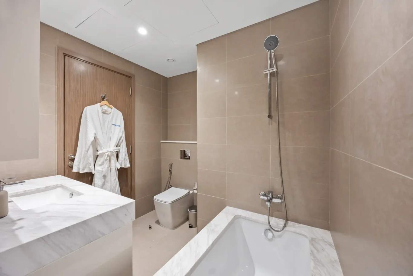 Modern beige-tiled bathroom with a walk-in shower, bathtub, toilet, and a white robe hanging on a wooden door