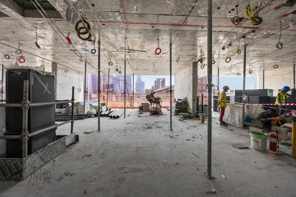 Interior of a high-rise under construction with exposed wiring and scaffolding, city skyline visible outside at the far end