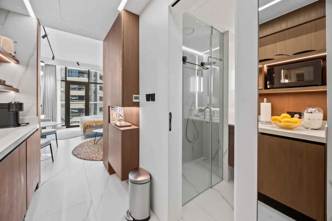 Modern apartment kitchen and bathroom area with wood cabinets, coffee station, and a glass-walled shower in the hallway.