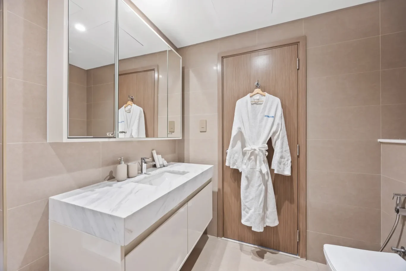 Modern bathroom with beige tile walls, a white marble countertop sink, and a wooden door with a white bathrobe hanging on a hook/door.