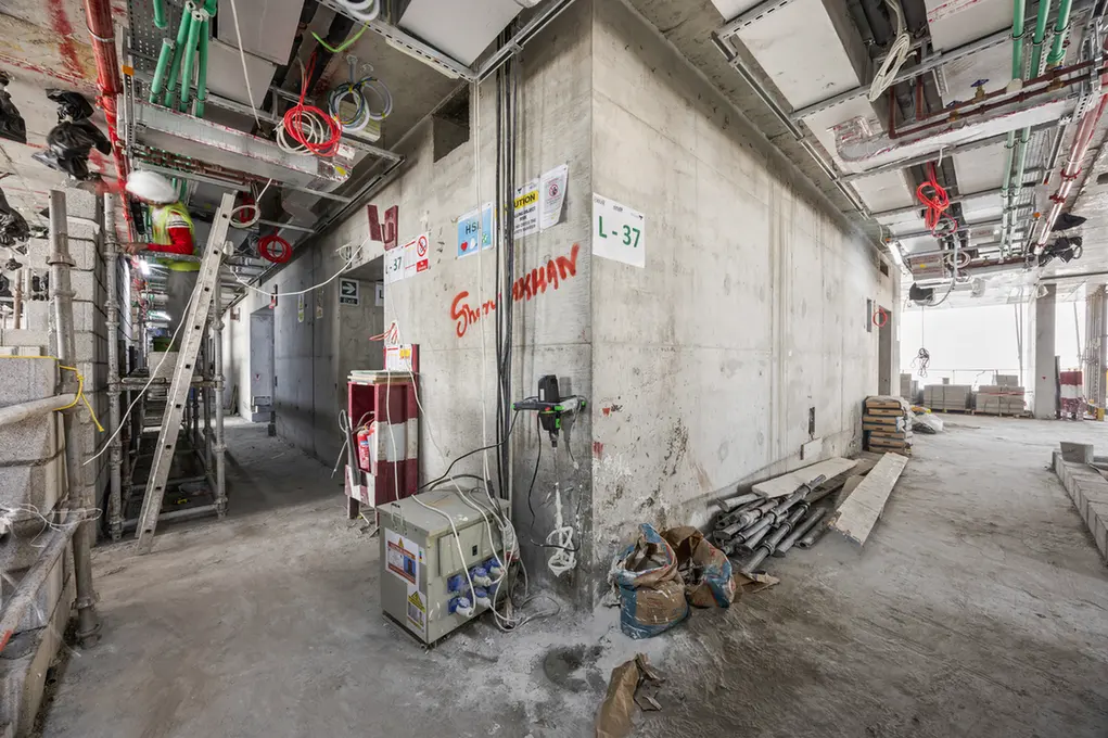 Construction site interior with scaffolding, exposed utilities, and a concrete pillar labeled L-37; dusty floor and work bags nearby still under construction.