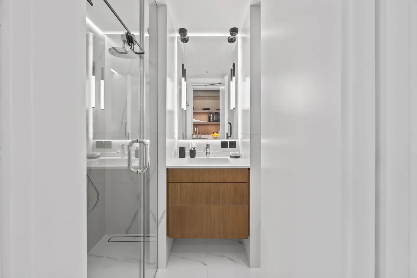 Narrow modern bathroom with glass shower, white marble floors, and a wooden vanity beneath a backlit mirror.