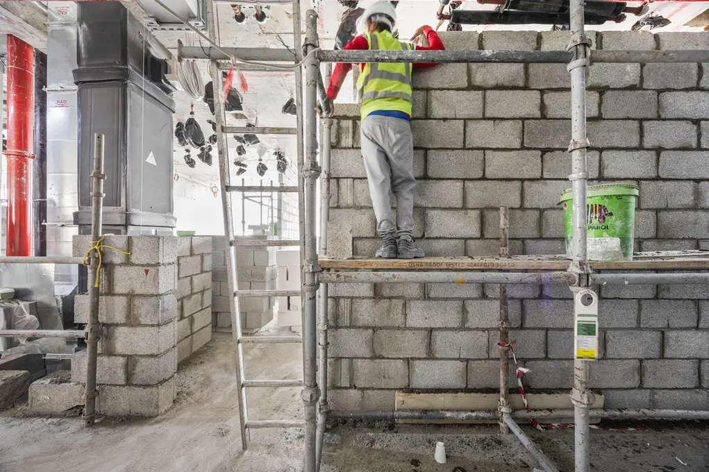Construction worker in a safety vest and helmet stands on scaffolding beside a partially built gray concrete block wall at a construction site, with a ladder nearby 1/2-bolts visible on the wall and piping above the workspace.