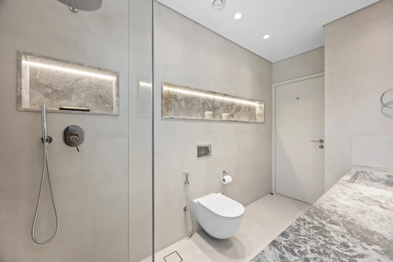 Minimalist bathroom featuring a glass shower enclosure, wall-mounted toilet, and marble accents with a recessed shelf of candles.