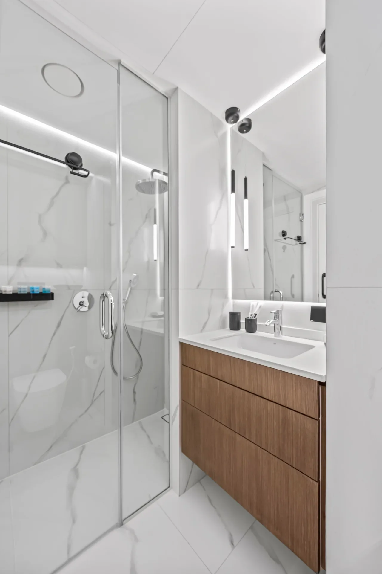 Modern white bathroom with glass shower enclosure, marble walls, and a wooden vanity under a backlit mirror.