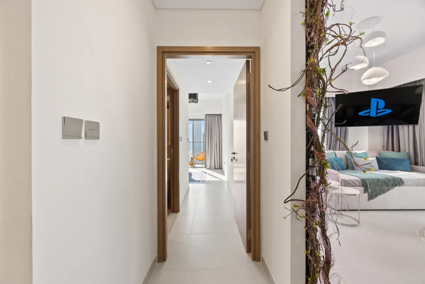 Bright hallway with a wooden doorway opening to a living area; white walls, light tiles, and a vine-wrapped divider beside a wall TV showing a blue PlayStation logo.