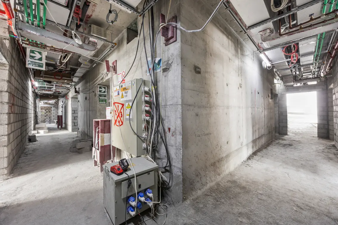 Construction hallway under renovation with exposed wiring, concrete walls, and electrical panels.