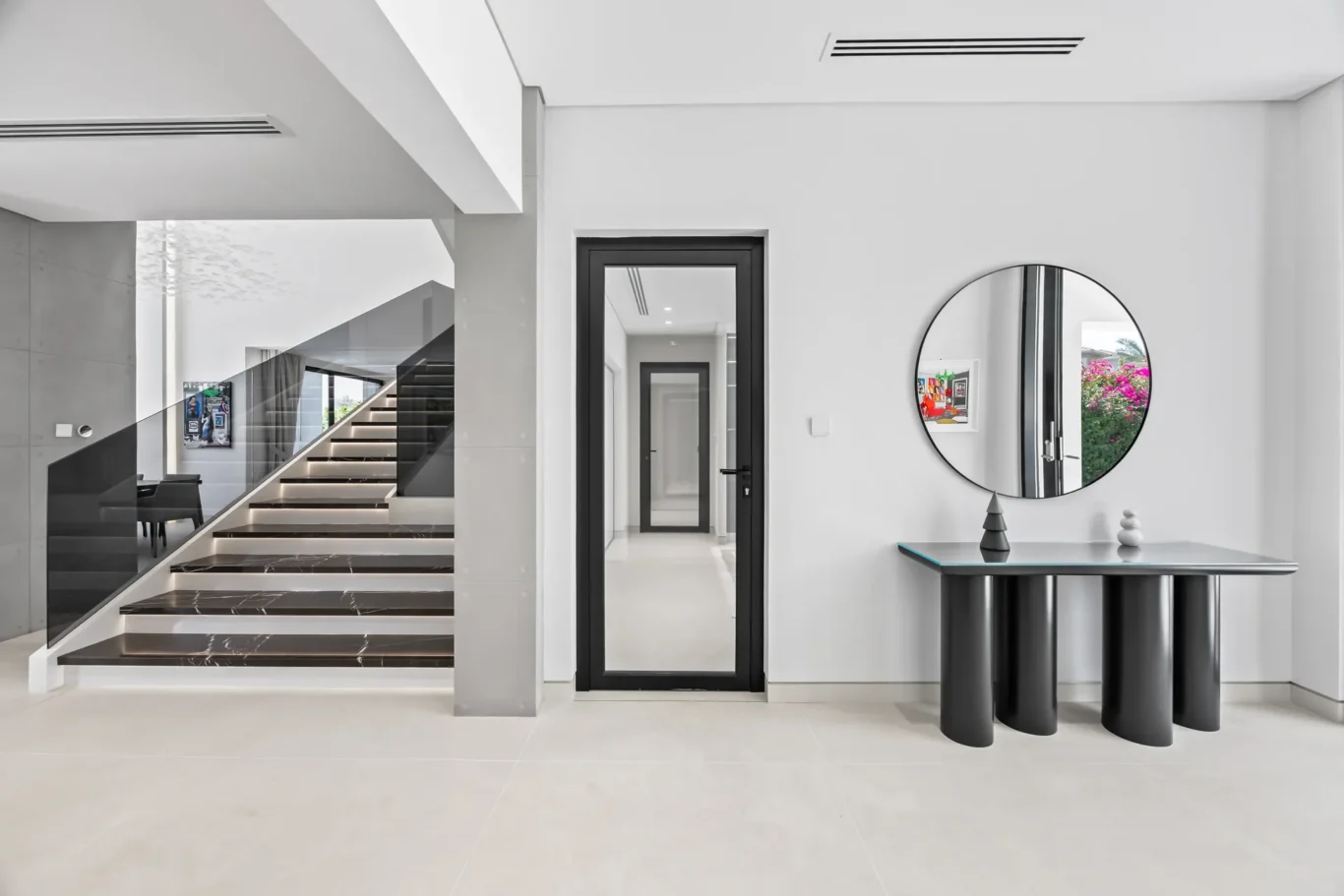 Contemporary white foyer with a black-framed glass door, round mirror, and sleek black console table; staircase on the left.