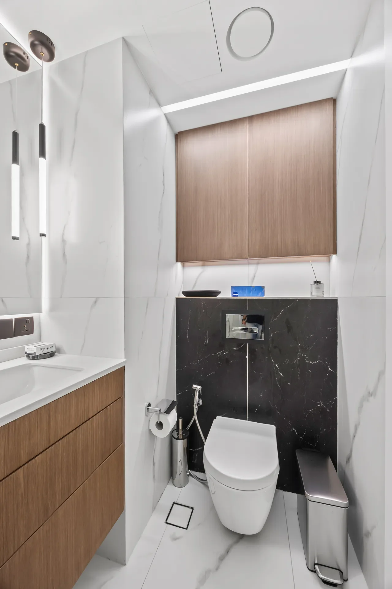 Modern bathroom with white marble walls, wooden vanity and upper cabinet, and a white toilet against a black marble panel.