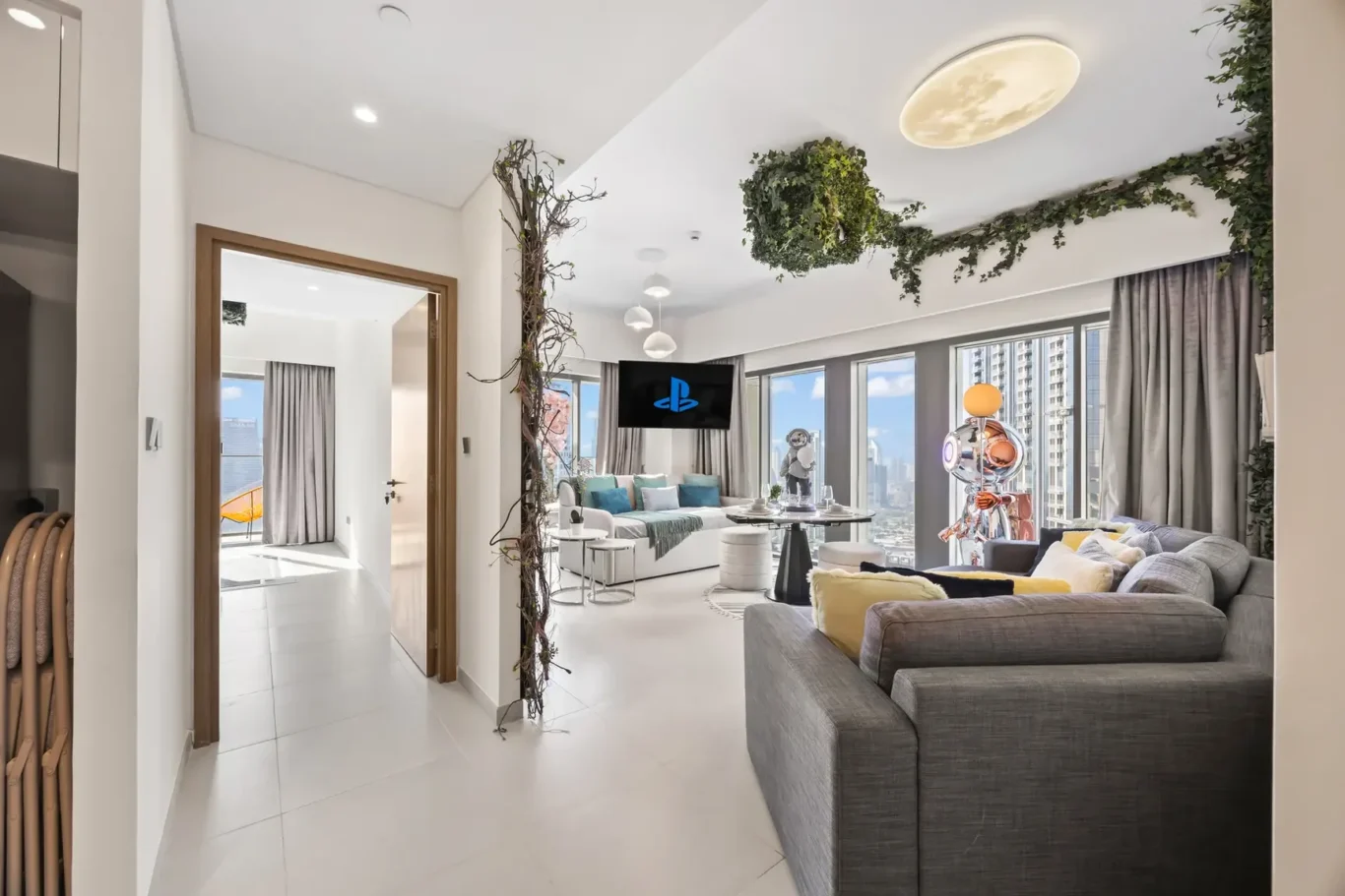 Bright open living room with a wall-mounted TV showing the PlayStation logo, white furniture, and large windows overlooking the city.