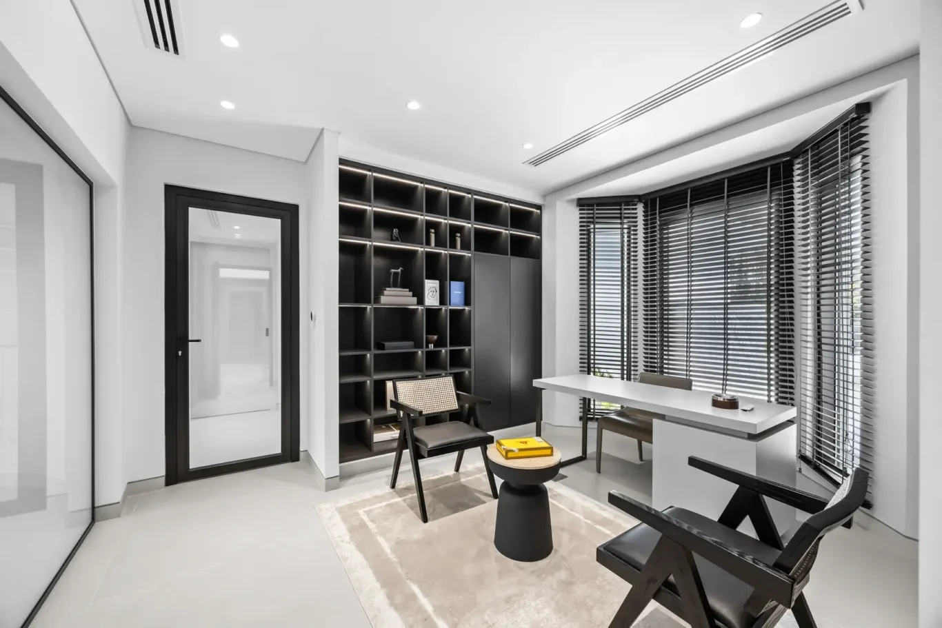 Bright minimalist office with a black modular shelving unit, white desk, and large sunlit windows with blinds.