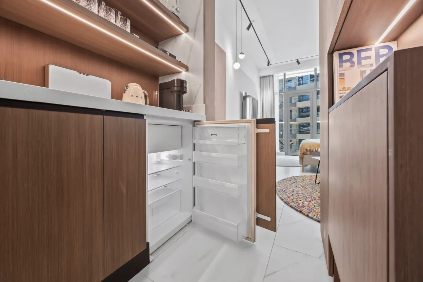 Open refrigerator in a modern kitchen with wood cabinets and a marble countertop, leading to a bright living area with a rug and large windows.