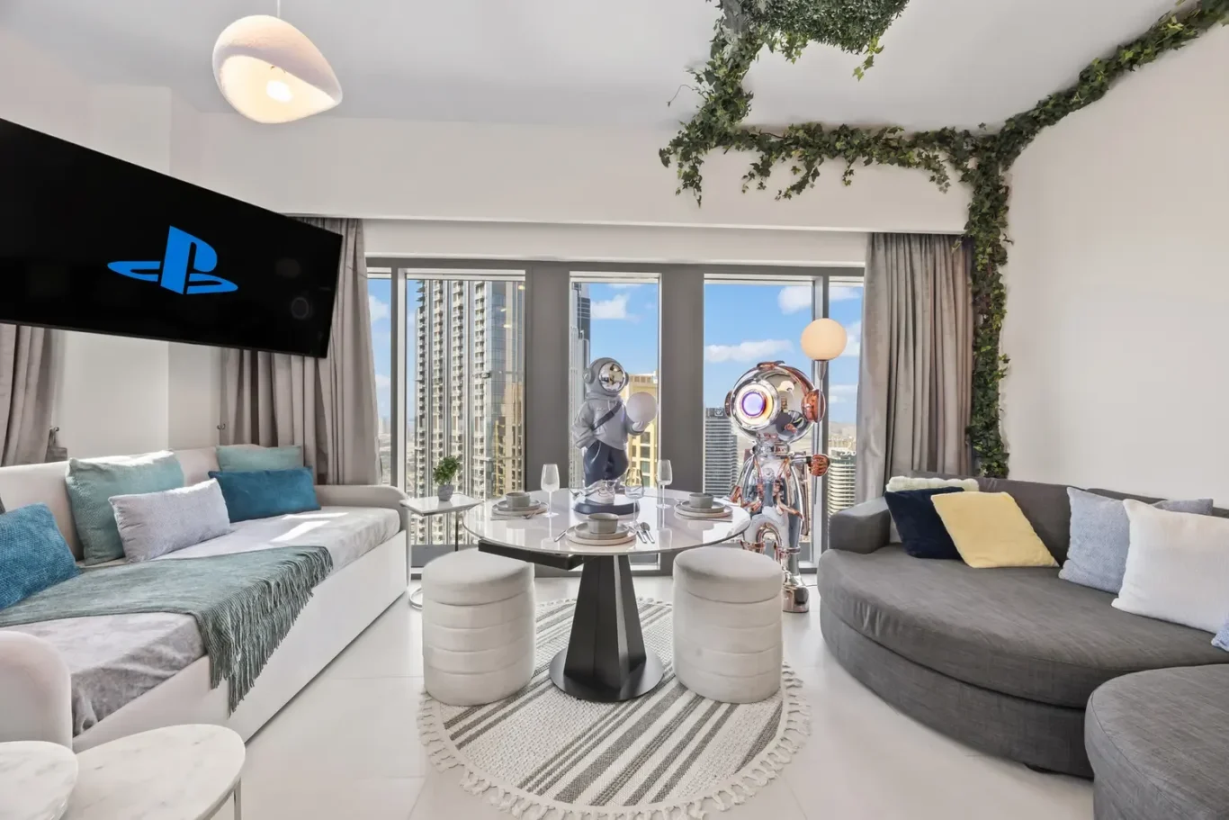 Modern living room with large windows showing a city skyline, white/gray furniture, and ivy decor along the ceiling. Two astronaut-like figures stand by a round glass table set for four.
