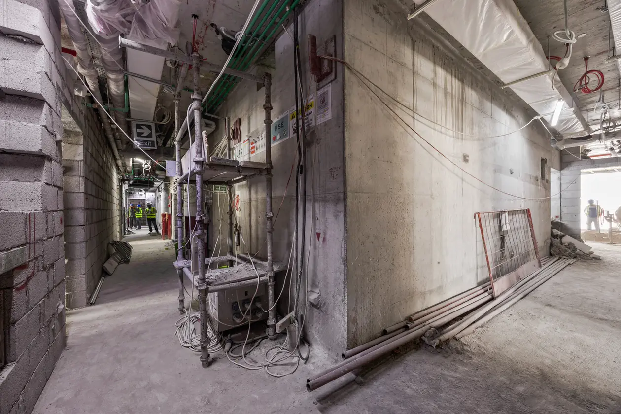 Narrow construction hallway with scaffolding, exposed pipes, and cables; workers in high-visibility vests in the distance.