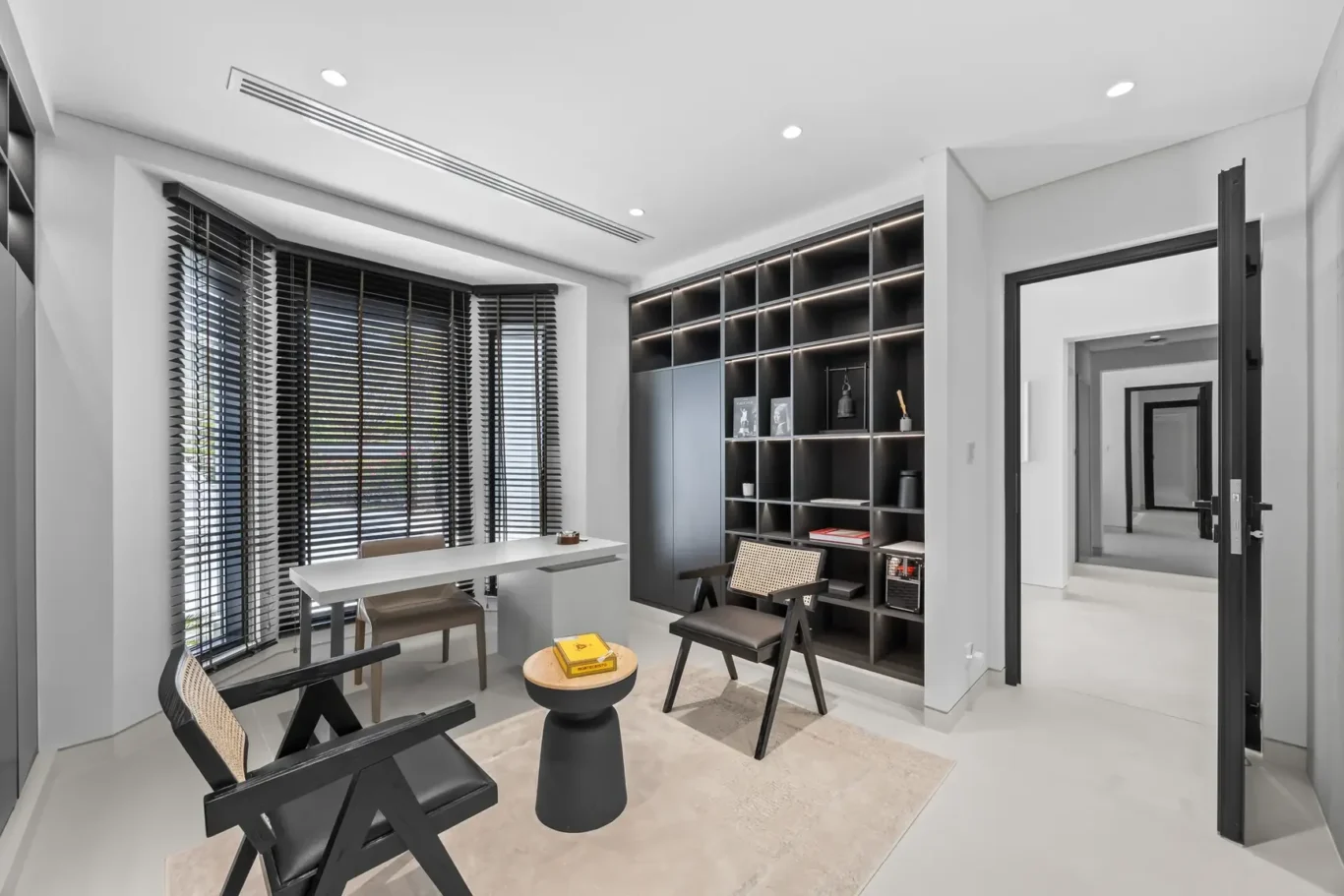 Modern home office with a white desk by large windows with black blinds and a built-in dark shelving unit on the right side.