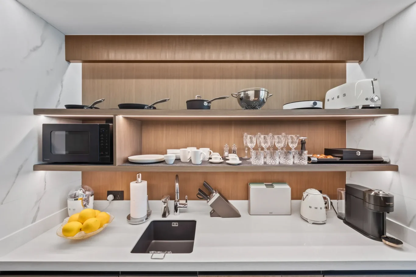 Open kitchen shelf with cookware, glassware, and small appliances including a microwave and a SMEG toaster on a wooden backdrop.