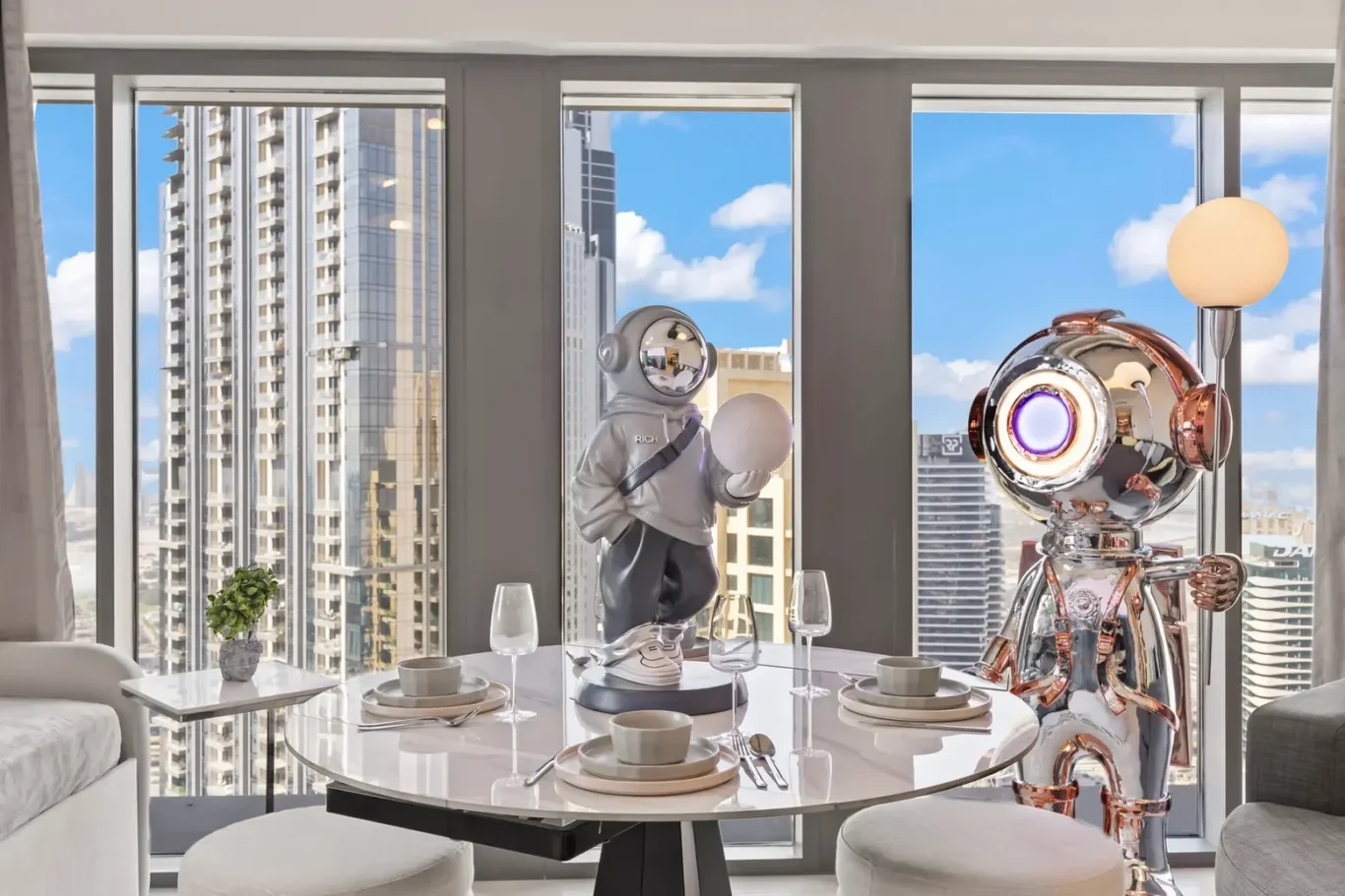 Two robots stand beside a round dining table set for four in a bright city apartment with a skyline view outside the windows, and a small plant on a side table.