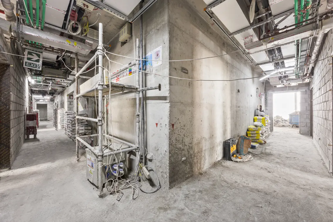Indoor construction site hallway with scaffolding, exposed piping, and stacked material bags along concrete walls.