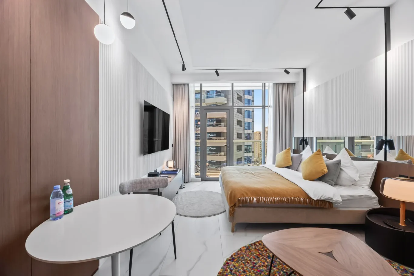 Bright hotel bedroom with a large window view, king bed with mustard throw pillows, and a white desk with water bottles nearby.