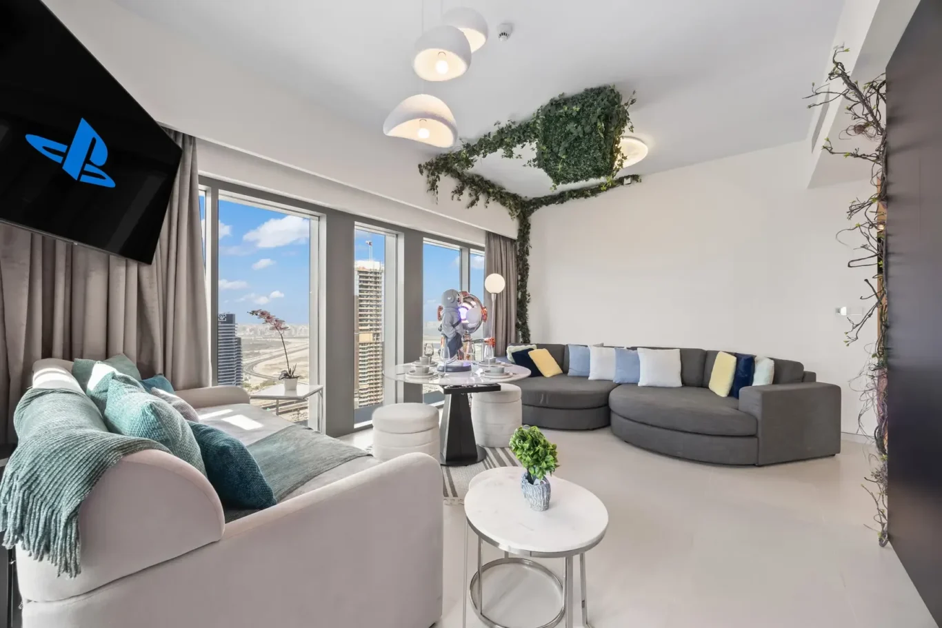 Bright modern living room with floor-to-ceiling windows showing cityscape, gray sectional sofa with colorful cushions, and white pendant lights over round coffee tables.