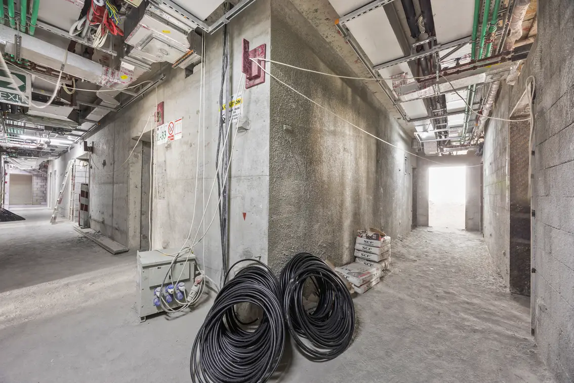 Unfinished concrete corridor in a building under construction with exposed wiring, pipes, and coiled black cables in the foreground.