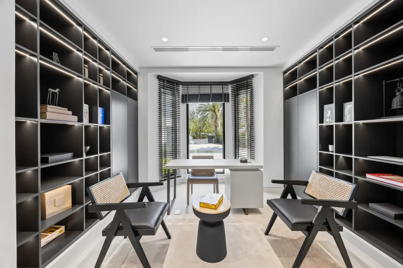 Modern home office with black built-in bookshelves, two black chairs, and a white desk facing a bay window with blinds.