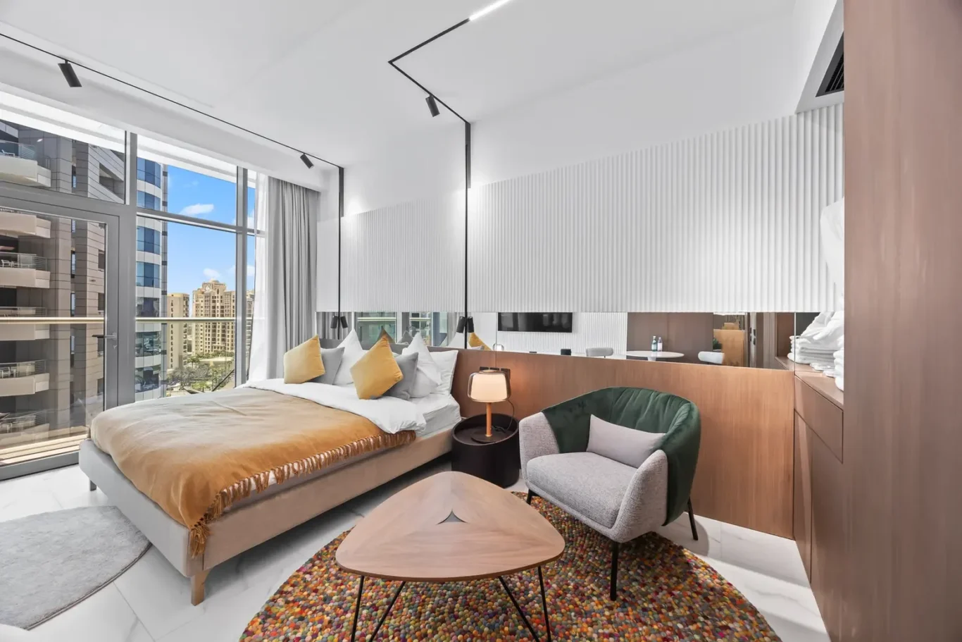 Modern hotel room with a low bed, mustard throw, gray and yellow pillows, and floor-to-ceiling windows showing city buildings—bright and airy.