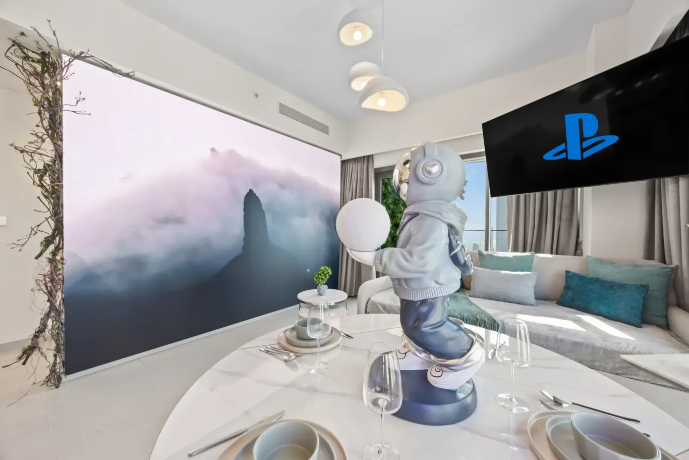 Modern living room with a misty mountain mural, an astronaut statue holding a white ball, and a round dining table set for dinner in the foreground.