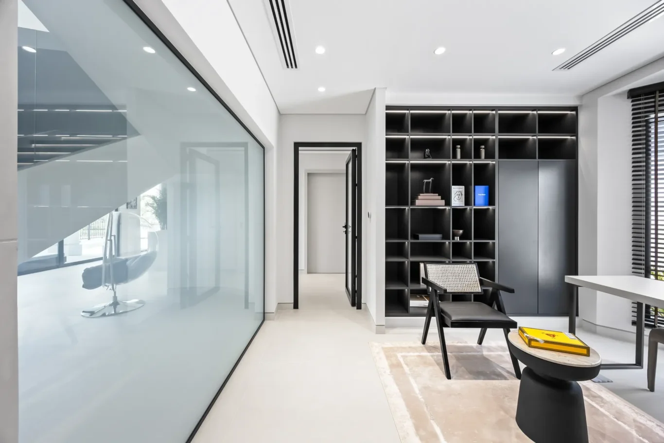 Modern office with glass partition, black shelving unit, and a black chair beside a round table with a yellow book.