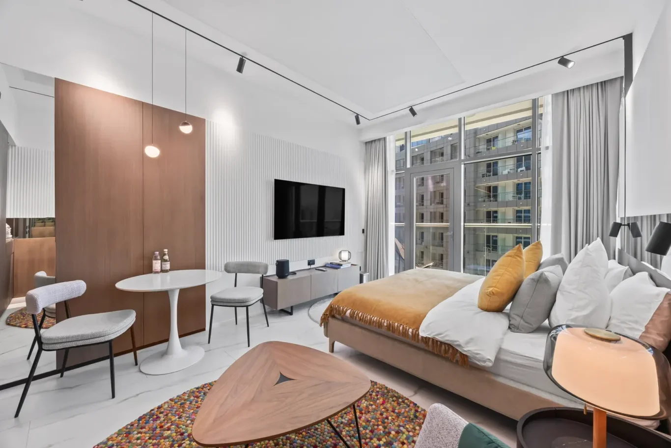 Modern hotel suite with a large bed, wall-mounted TV, seating area, and expansive windows overlooking the city.