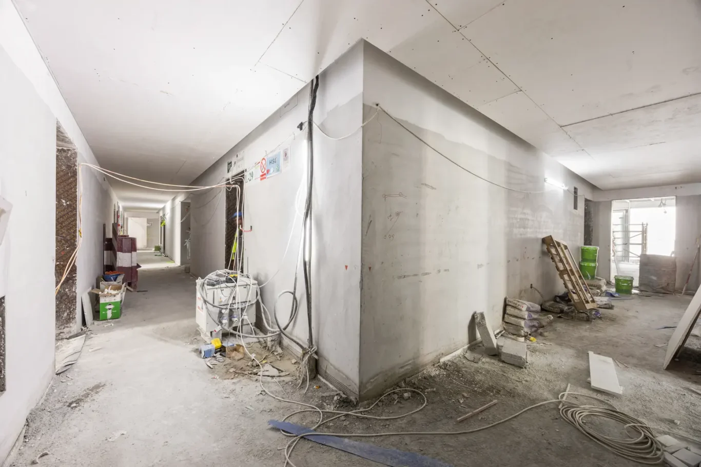 Construction site hallway with exposed wiring, concrete walls, and scattered debris and tools on the floor.