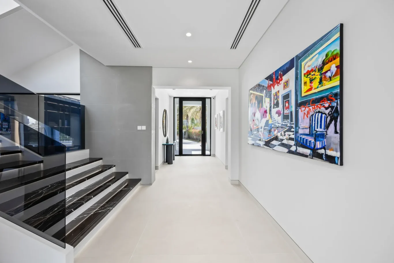Bright modern hallway with a glass-railed staircase on the left, white walls, and a large colorful painting on the right wall, leading to a glass door opening to a garden.