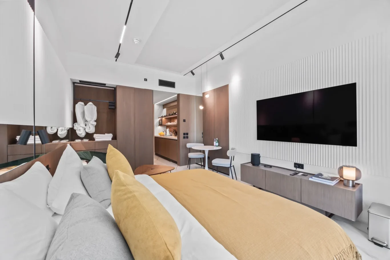 Contemporary hotel suite living area with a bed in the foreground, mustard pillows, and a large flat-screen TV on a white slatted wall.