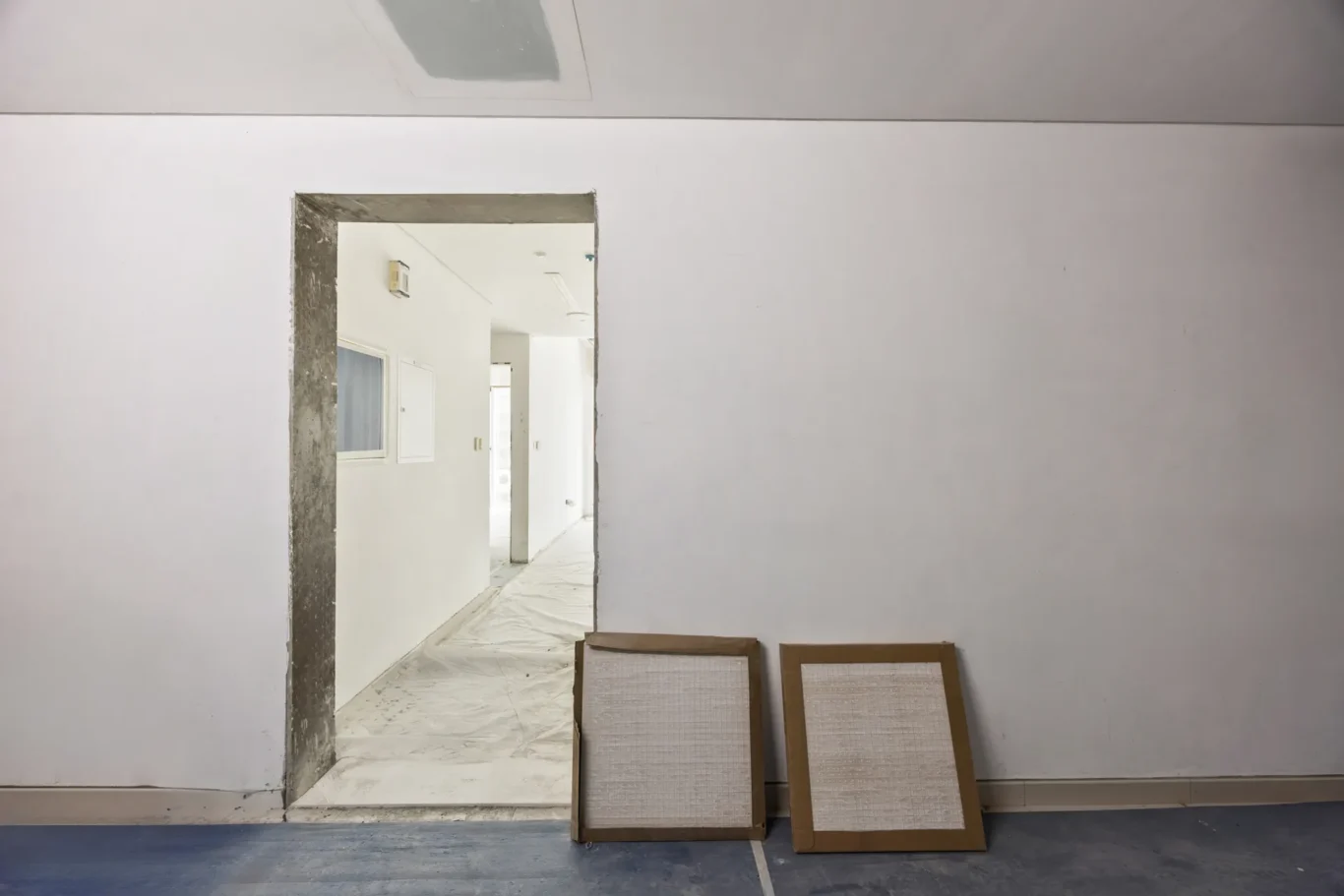 Interior of a building under renovation: open doorway to a white-walled corridor, with two large framed panels leaning against the wall in the foreground.