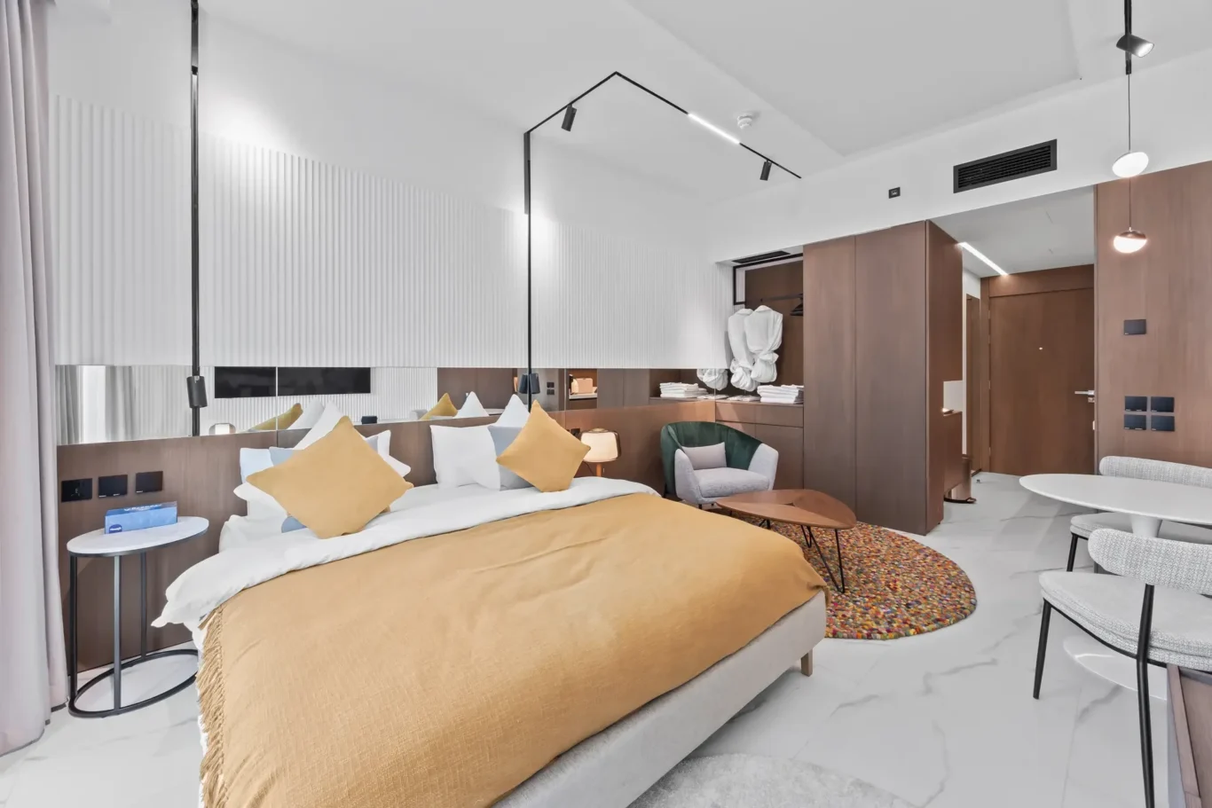 Modern hotel bedroom with a mustard duvet, white linens, and yellow cushions; wood wardrobe and seating area nearby.