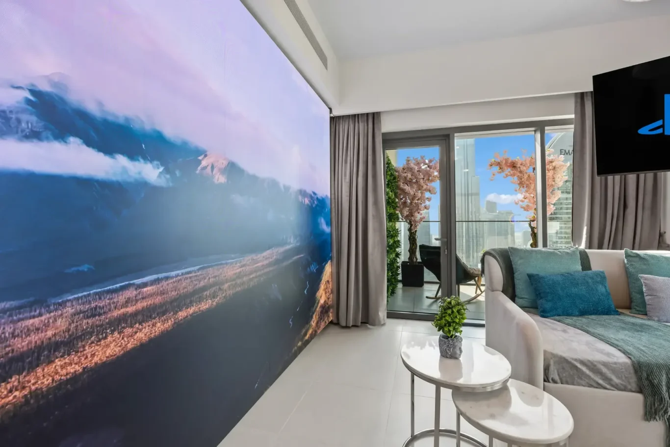 Living room with a large landscape mural on the wall, glass doors to a balcony, and a sofa with teal cushions.