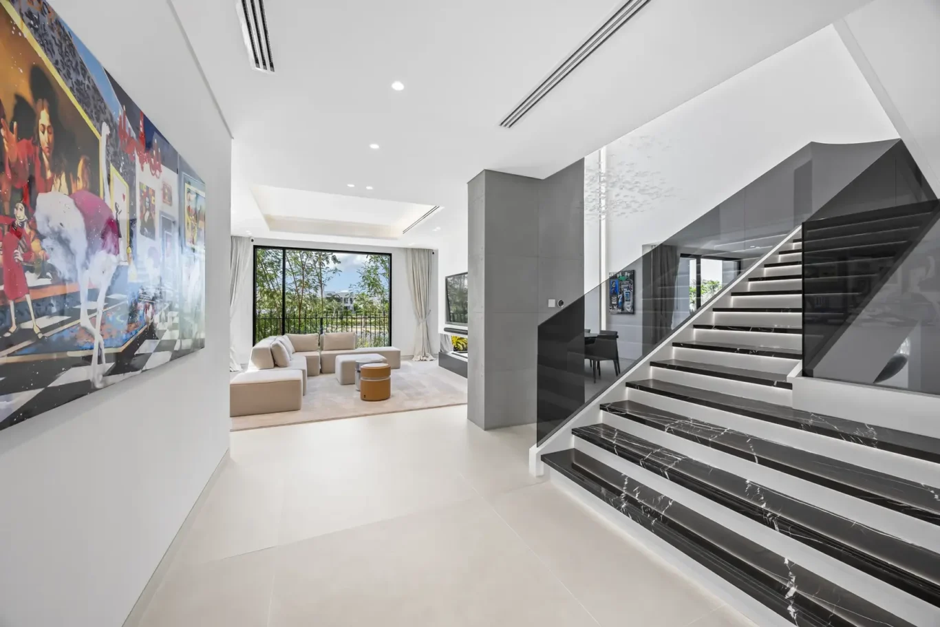 Bright modern living area with a beige sectional, a large wall art piece, and a glass-railed marble staircase to the right.