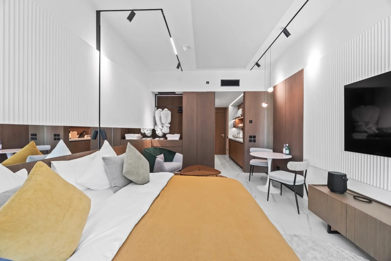 Contemporary hotel suite living area with a large bed in the foreground, mustard bedding, and a wood-accented layout.