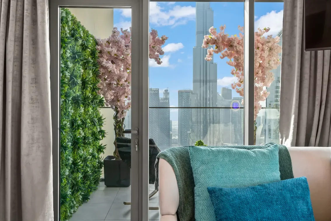 Living room with glass doors opening to a balcony, a green plant wall outside, pink blossoms, and a city skyline in the background; teal cushions on a light sofa.