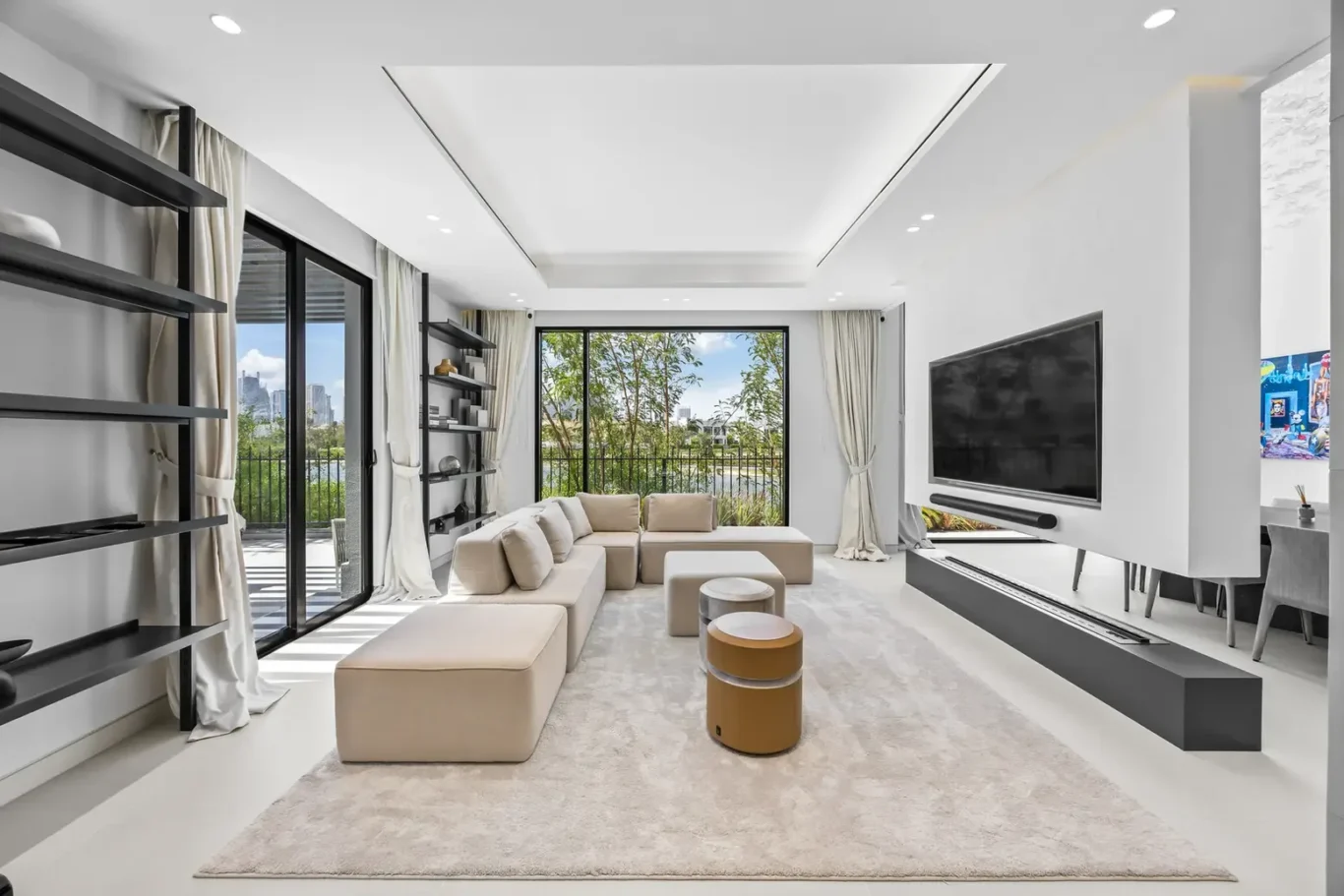 Bright modern living room with a beige sectional sofa, large windows, and a wall-mounted TV overlooking a balcony with greenery.