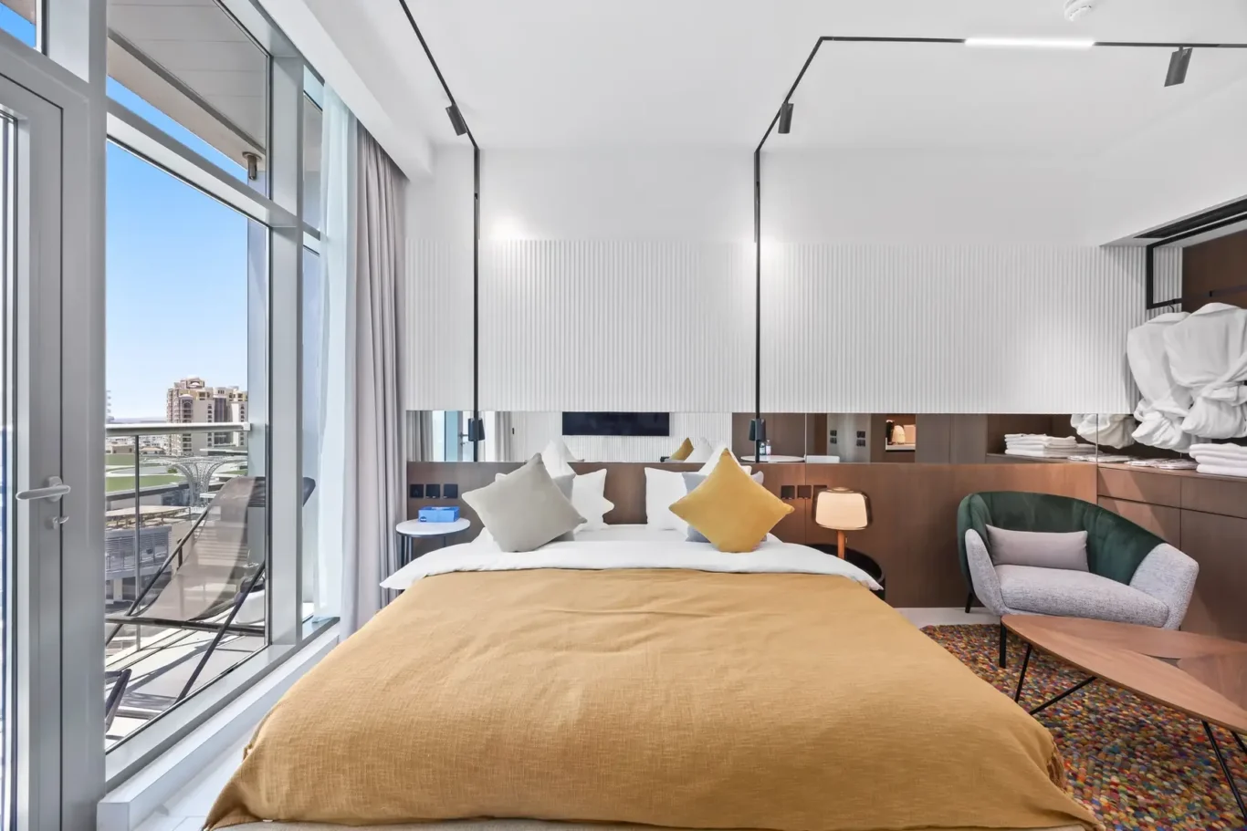 Bright hotel room with a large bed, mustard bedding, and colorful throw pillows, overlooking a balcony with city view through floor-to-ceiling glass doors.