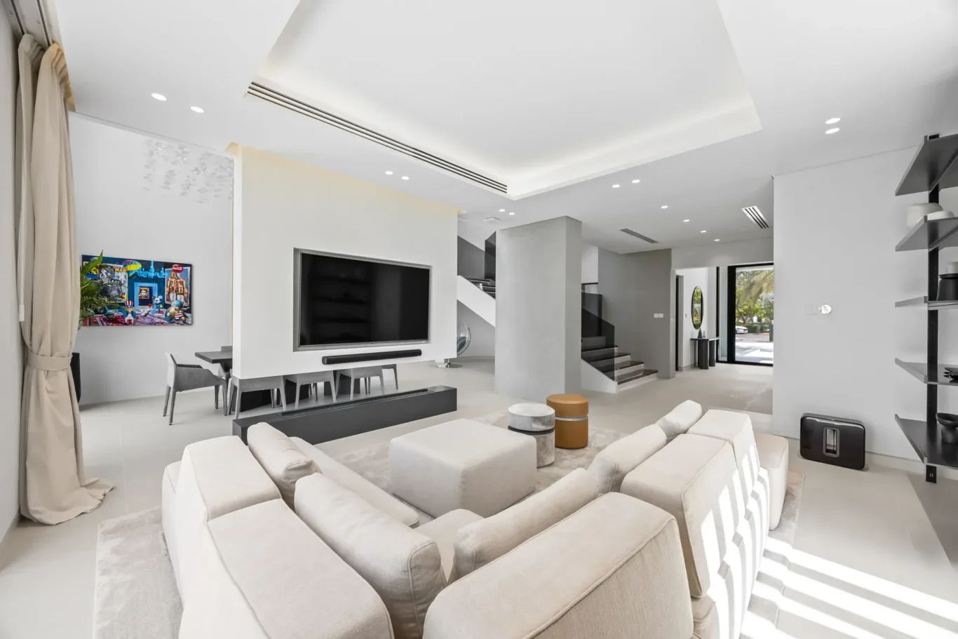 Open-concept living room with white seating around a wall-mounted TV and a sleek media console, stairs in the background, and bright natural light.