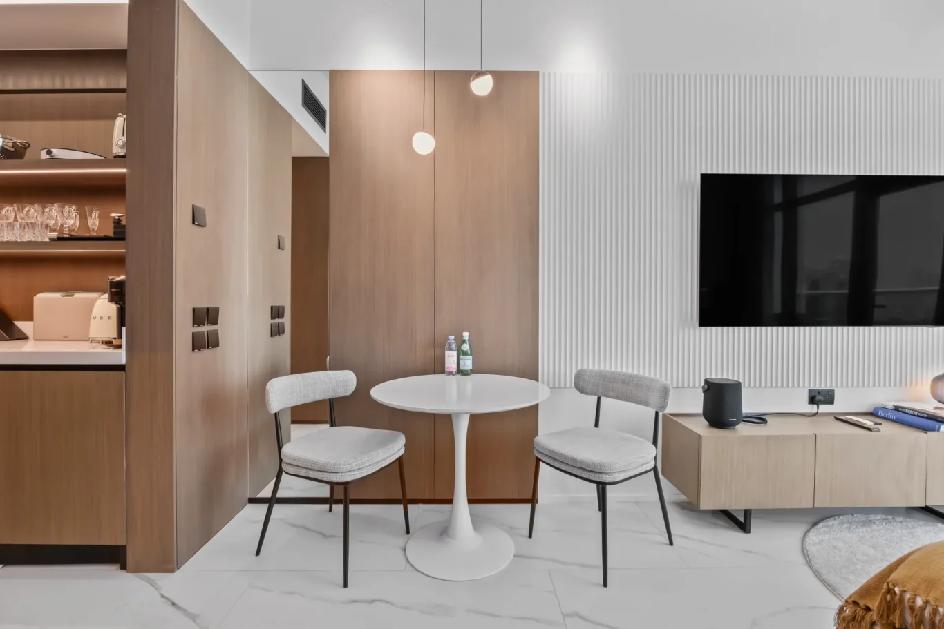 Round white pedestal table with four gray upholstered chairs in a modern kitchen-living area; wood cabinetry, pendant lights, and a wall-mounted TV on a ribbed wall.