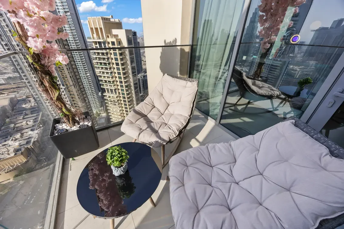 Balcony with glass railing overlooking city skyscrapers; two tufted lounge chairs and a round black table with a plant.