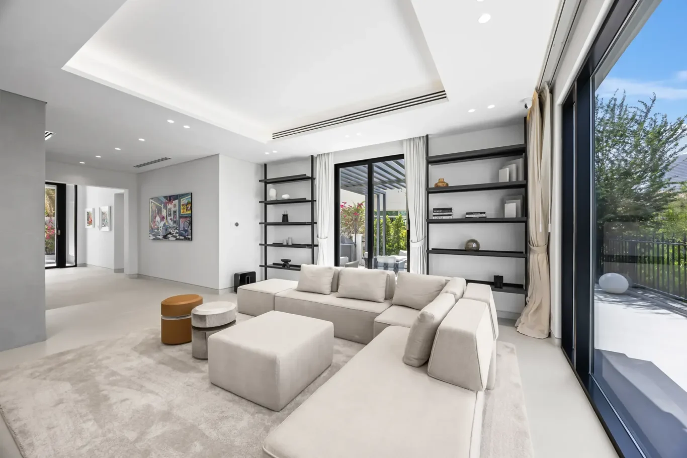 Bright, minimalist living room with a light gray sectional sofa, round ottomans, and black open shelves by large glass doors to an outdoor patio