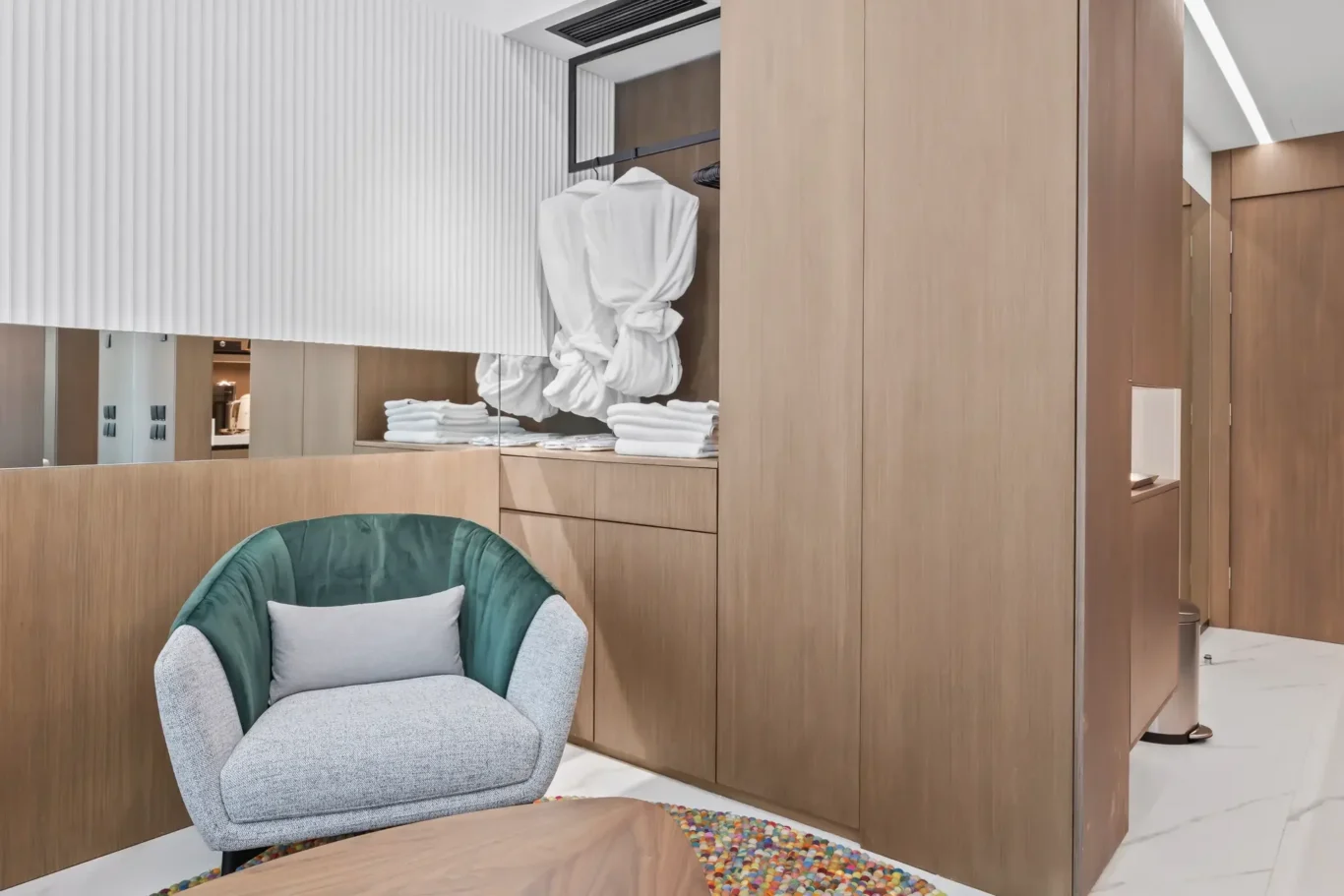 Open wardrobe area with a white bathrobe hanging and stacks of folded towels on wooden shelves, a green upholstered chair in the foreground, and light wood panels creating a modern, luxurious dressing space.