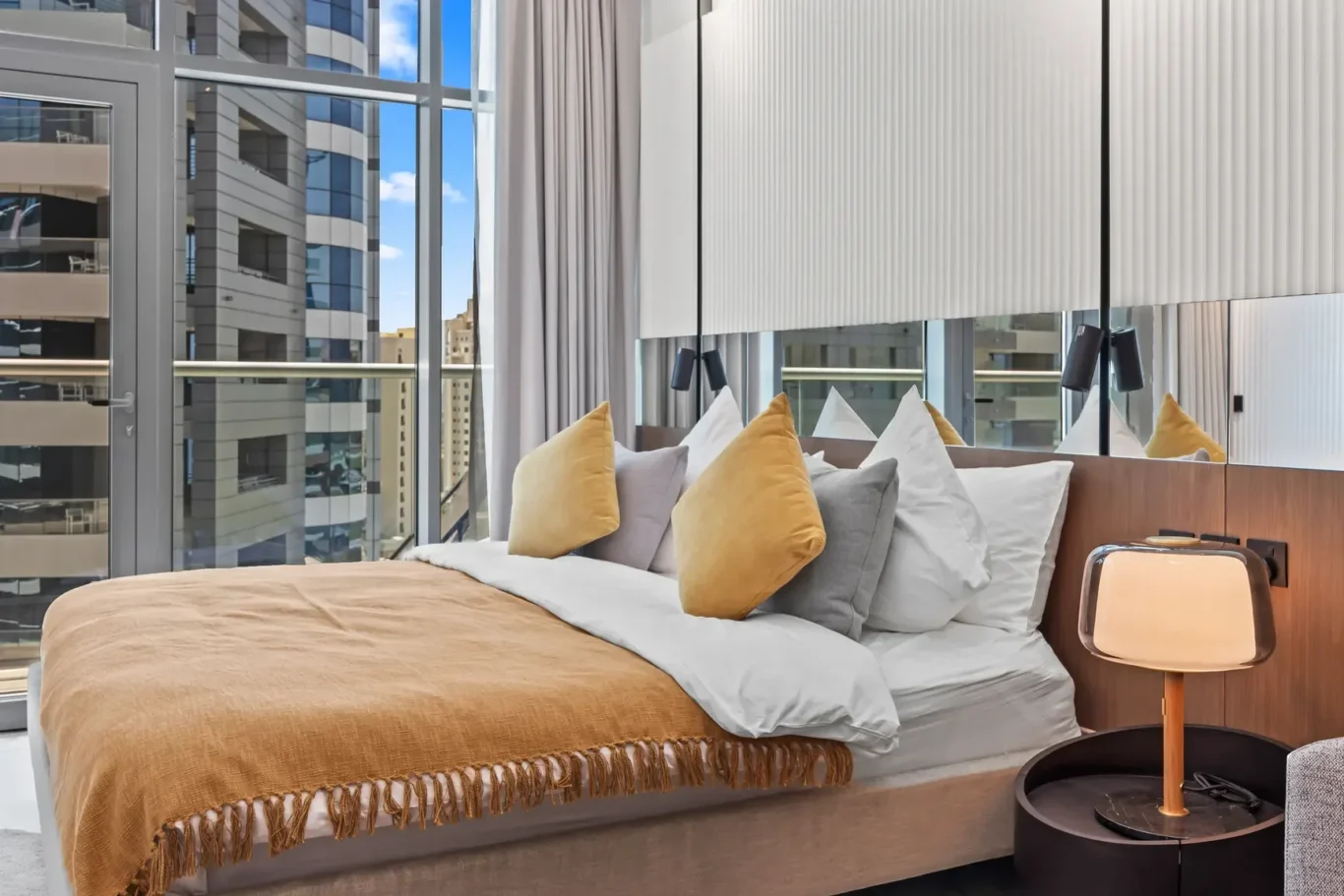 Hotel bedroom with large windows and city view, bed made with beige throw and multiple pillows.