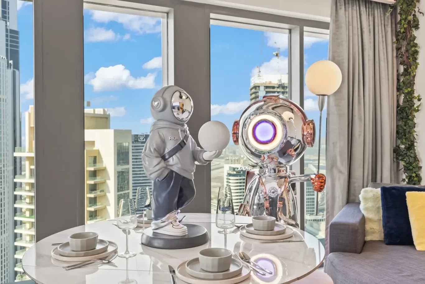 Two shiny robots share a meal at a glass dining table in a high-rise apartment, with a city skyline outside the windows.